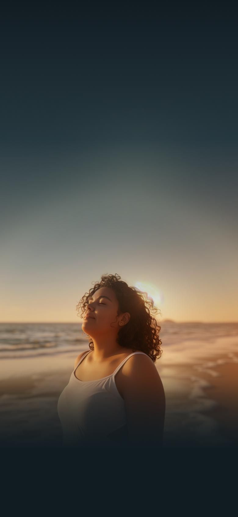 A woman with curly hair stands on a beach at sunset, eyes closed and serene.