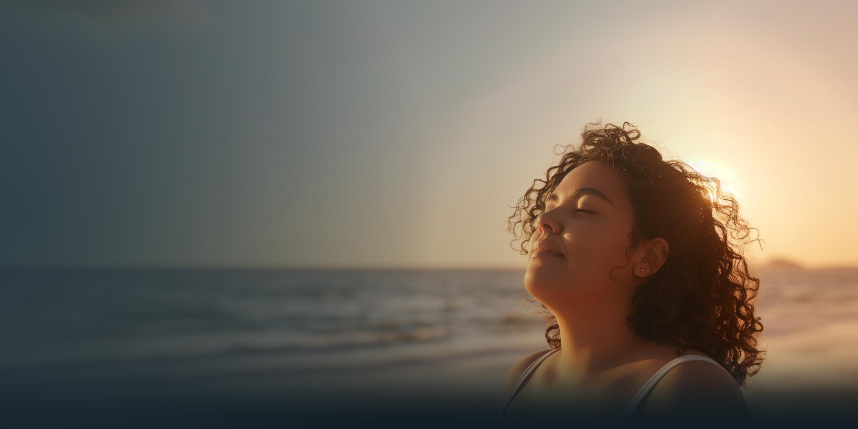 A woman with curly hair stands on a beach at sunset, eyes closed and serene.