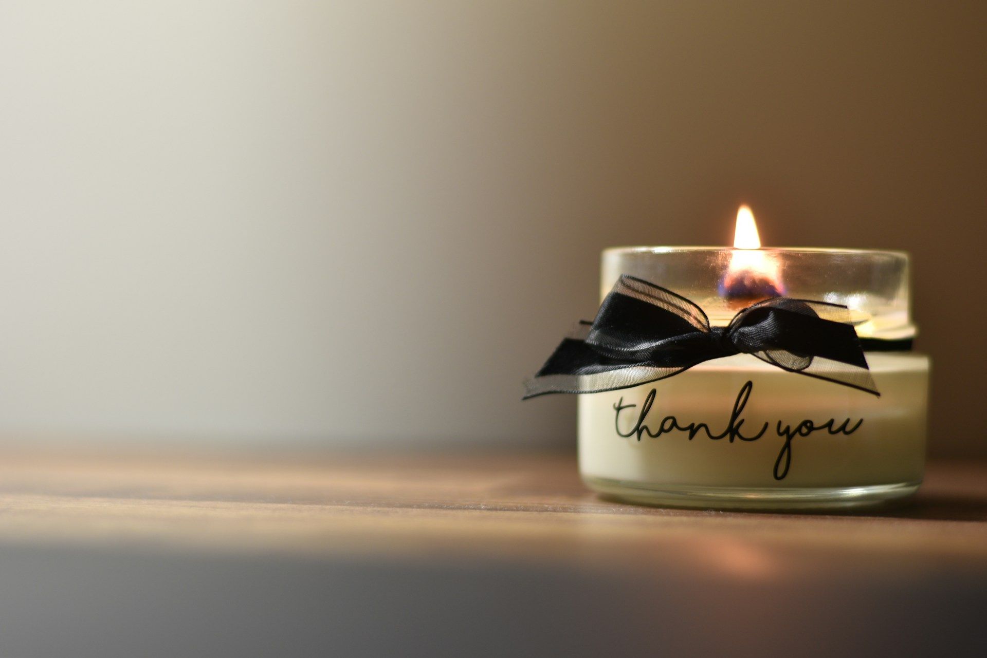 A lit candle in a glass jar with a black ribbon and “thank you” written on it, placed on a wooden surface with a soft gradient background.