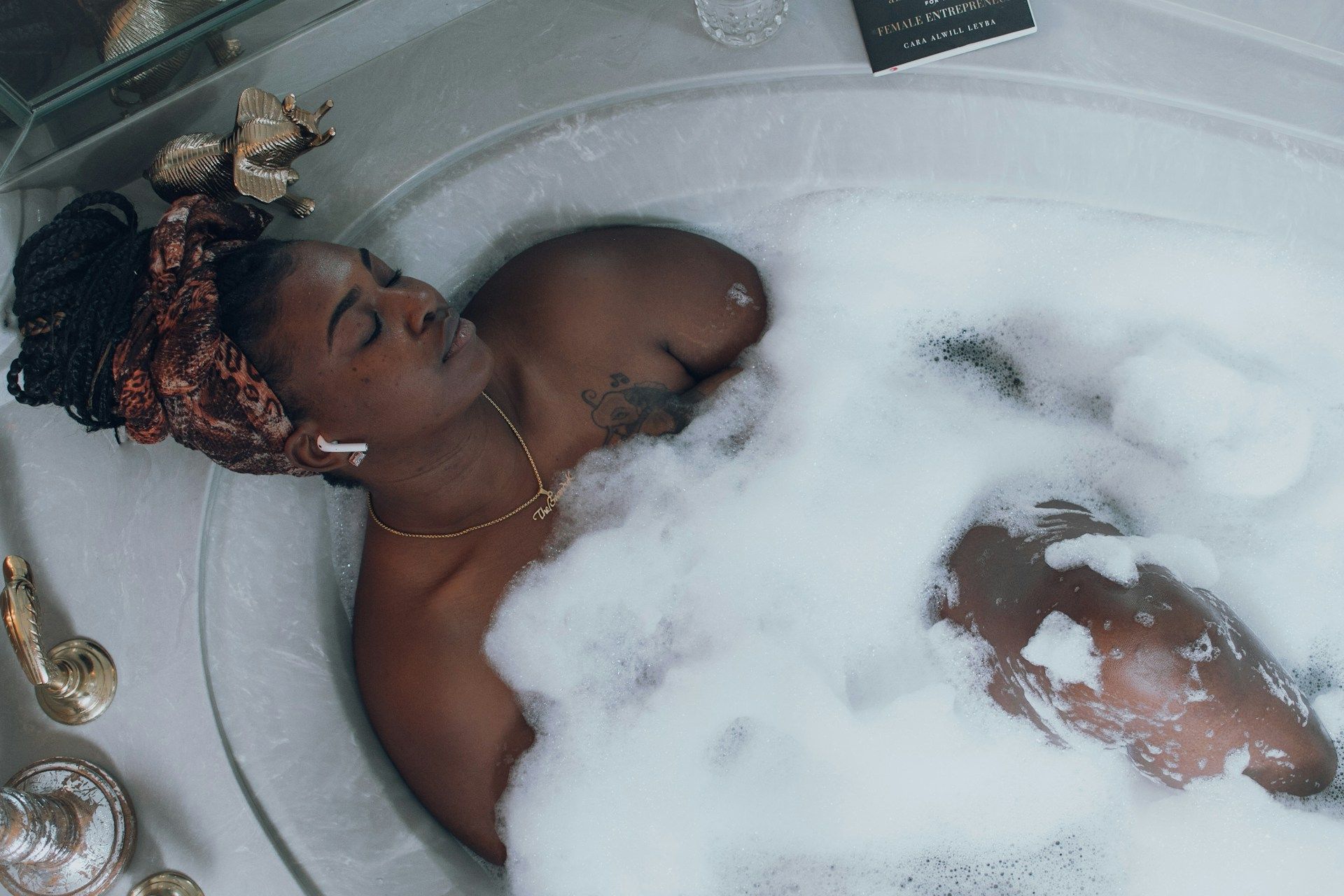 A woman with a headscarf and AirPods is relaxing in a bubble bath with her eyes closed