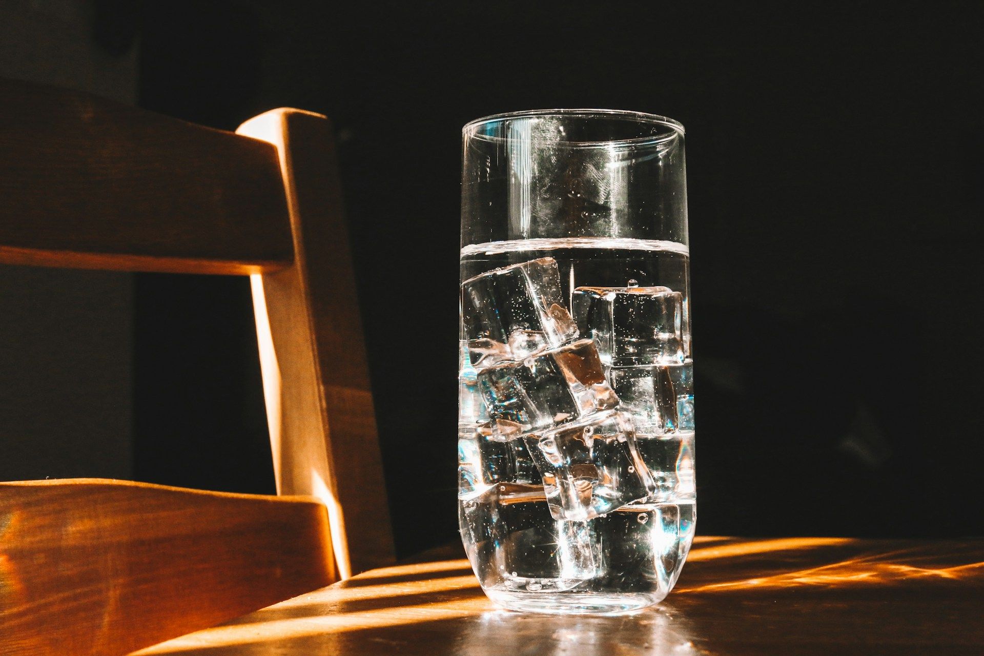 A tall glass of ice water on a wooden surface, illuminated by sunlight with a dark background.