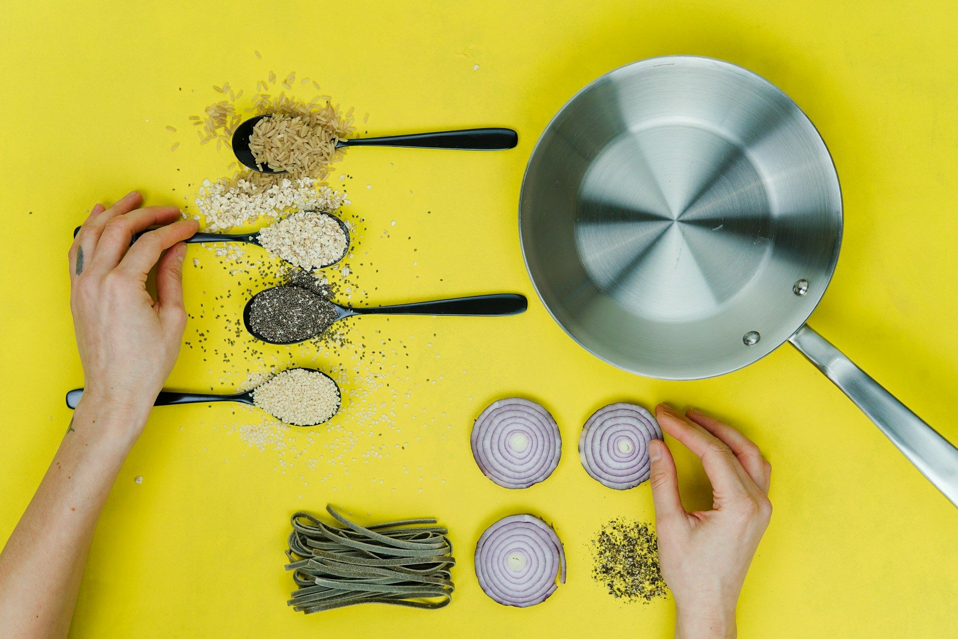 Hands preparing healthy ingredients: various grains on spoons, red onion slices, and a cooking pot on a bright yellow background