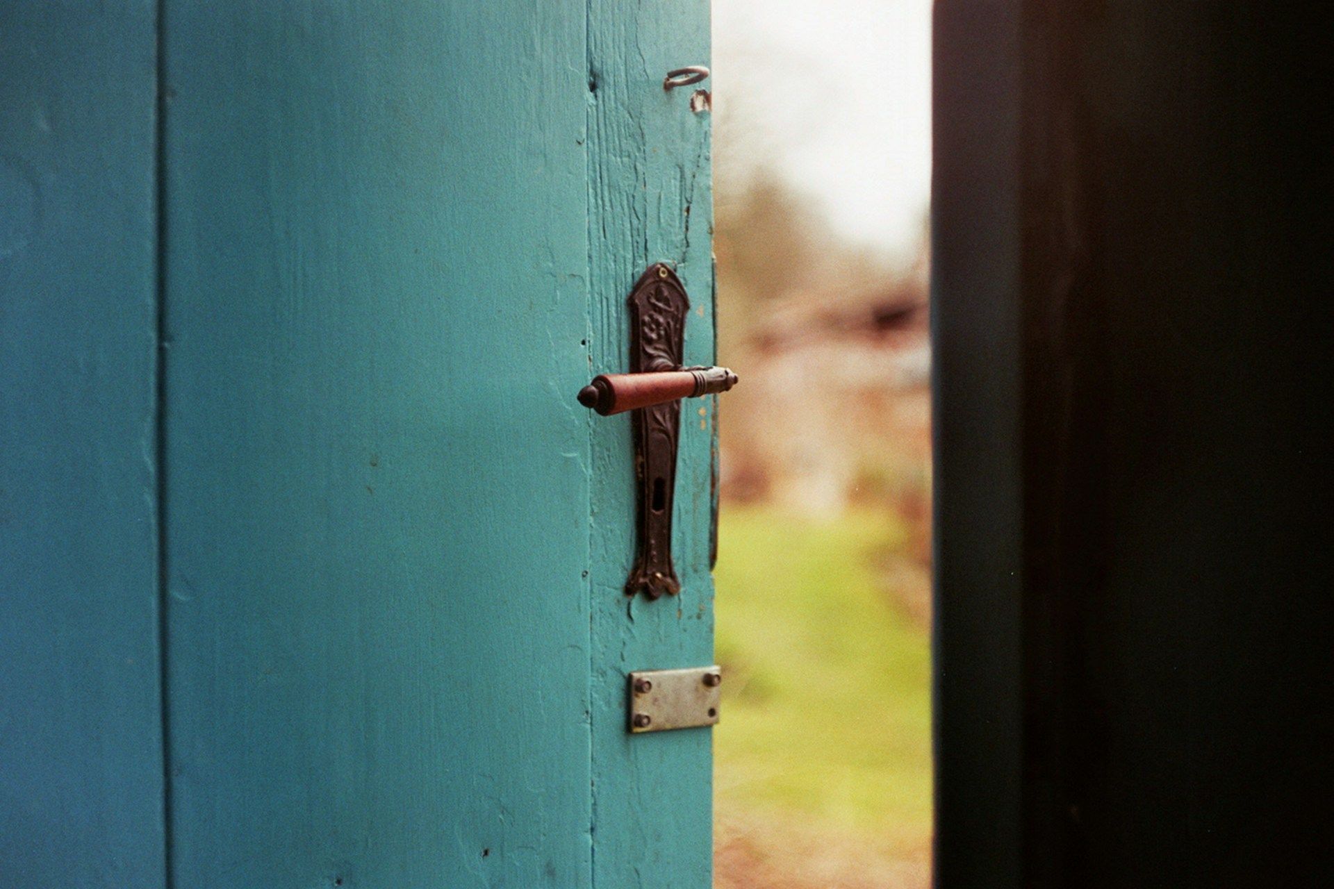 A partially open turquoise door with a vintage metal handle, leading to a blurred garden background