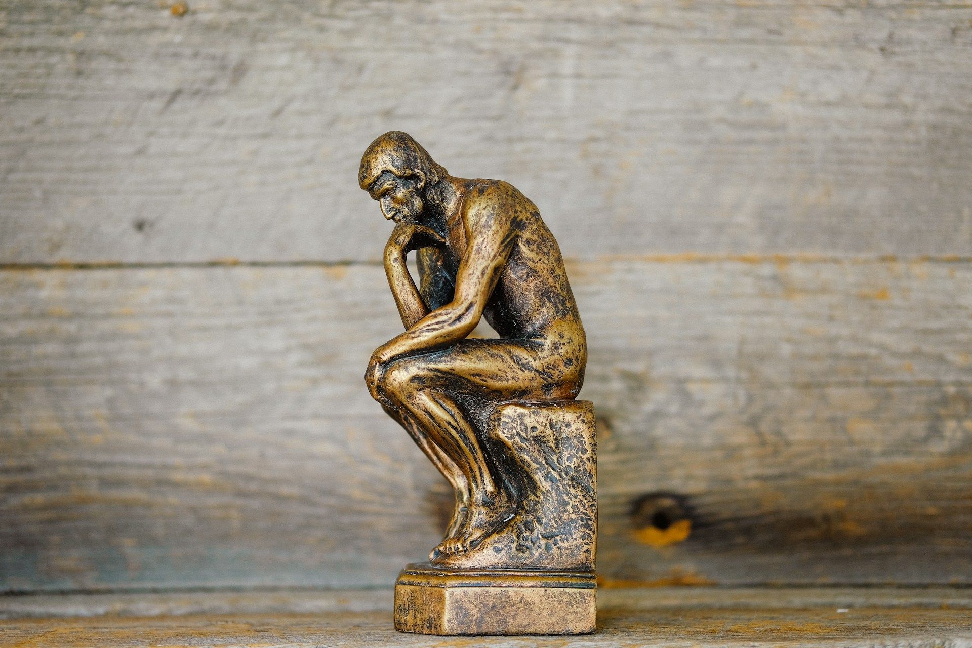 A small bronze statue of a man in a contemplative pose, seated on a square pedestal with his chin resting on his hand, resembling Auguste Rodin’s “The Thinker,” against a rustic wooden background